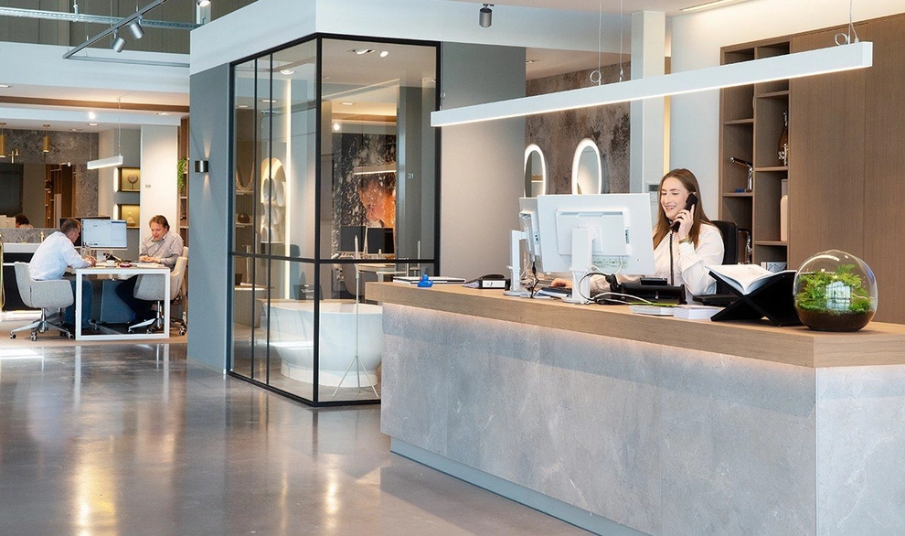 Une femme souriante derrière le comptoir d’accueil dans un bureau moderne avec une expérience olfactive subtile créant une atmosphère chaleureuse et accueillante.